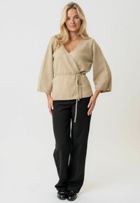 Beiger Wickelpullover mit langen, weiten Ärmeln und einem gebundenen Taillenband. Der Stoff wirkt weich und strukturiert, kombiniert mit schwarzen, gerade geschnittenen Hosen.