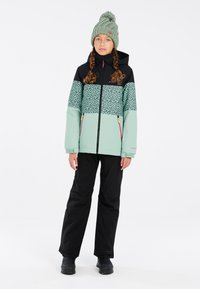 Giacca verde menta e nera con motivo a pois, abbinata a pantaloni neri e un berretto di maglia verde; presenta cerniere e una vestibilità rilassata.