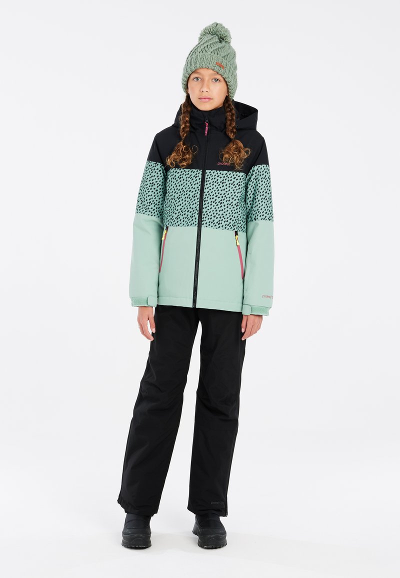 Giacca verde menta e nera con motivo a pois, abbinata a pantaloni neri e un berretto di maglia verde; presenta cerniere e una vestibilità rilassata.