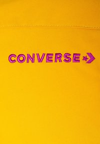 Żółty materiał z fioletowym haftowanym napisem "CONVERSE" i logo gwiazdy-zębatki poniżej przeszytej szwy.