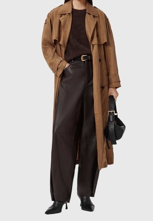 Femme portant un trench-coat marron, un pull marron foncé, un pantalon large en cuir noir, une ceinture noire, des bottes pointues noires et tenant un sac à main noir.