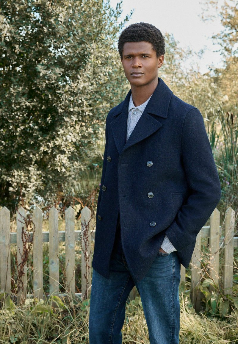 Marineblauer, doppelt geknöpfter Peacoat aus strukturiertem Wollstoff, mit großen Knöpfen, getragen über einem hellgrauen Hemd mit Kragen und blauen Jeans.