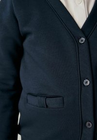 Gros plan d'un cardigan bleu marine foncé avec des boutons, un petit détail de ceinture en tissu sur la poche, porté par-dessus une chemise blanche à col.