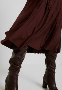 Brun plisserad kjol med flytande drapering, kombinerad med knähöga bruna läderstövlar med fyrkantig klack och synlig dragkedjedetalj.