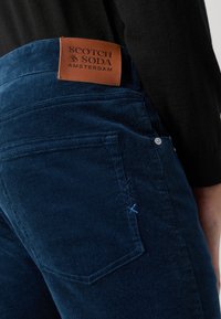 Temno modri kord hlače z rjavo usnjeno nalepko z logotipom blagovne znamke, dekorativnim šivanjem ob strani in prilegajočim dizajnom.