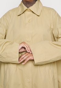 Beige, knappad regnrock med en stor, vikt krage, breda ärmar och sidofickor; tillverkad av ett slätt, lätt material.