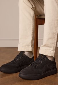 Baskets en daim noir avec une finition texturée, semelle en caoutchouc plate et œillets en métal, assorties à un pantalon et des chaussettes de couleur claire.