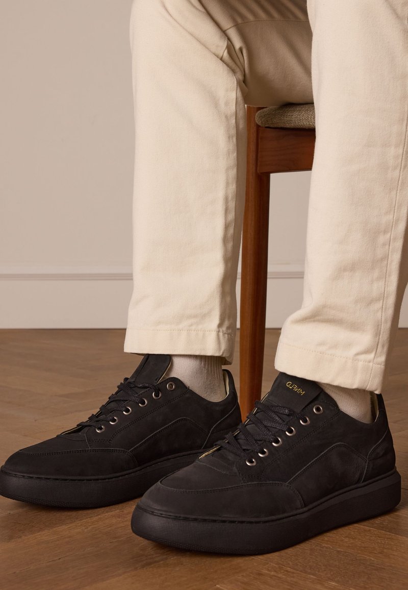 Baskets en daim noir avec une finition texturée, semelle en caoutchouc plate et œillets en métal, assorties à un pantalon et des chaussettes de couleur claire.