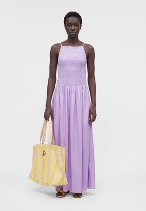 Žena v levanduľových maxi šatách bez rukávov drží veľkú žltú pruhovanú tašku, stojaca na svetlom jednoliatom pozadí.