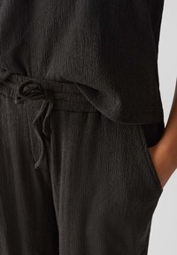 Tenue noire texturée avec une coupe ample, une taille élastique avec cordon de serrage et des poches latérales. Le matériau a une apparence froissée.