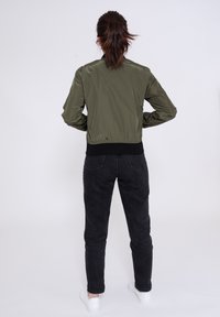 Giacca bomber verde oliva con una texture liscia, polsini arricciati e orlo a coste neri, indossata con jeans scuri e sneakers bianche.