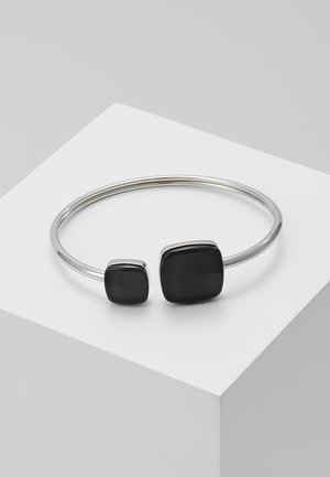 Hopeinen avoin rannerengas, jossa on kaksi mustaa neliönmuotoista kiveä eri kokoisina, esiteltynä valkoisella jalustalla.