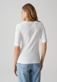 Frau mit langen Haaren steht mit dem Rücken zur Kamera, trägt ein weißes Oberteil mit Ellenbogen-langen Ärmeln und hellblaue Jeans vor einem einfarbigen hellen Hintergrund.