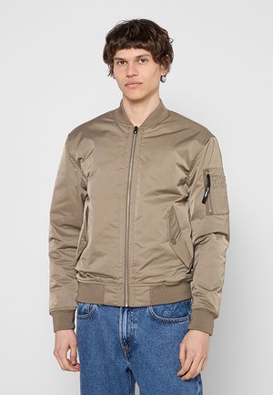 Junger Mann trägt eine beige Bomberjacke mit Reißverschluss und blaue Jeans, steht vor einem einfachen hellen Hintergrund.