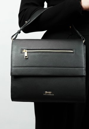 Sac à main en cuir noir avec un rabat supérieur, une poche zippée à l'avant et des finitions en métal argenté. Texture lisse avec un logo embossé.