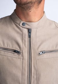 Beige suède jas met een hoge kraag, centrale ritssluiting en twee ritszakken op de borst. Voorzien van zwarte hardware en een gladde textuur.