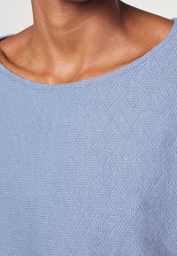 Ljusblå tröja med vid halsringning och en texturerad fiskbensmönster, som visar ett mjukt, vävt tyg nära huden.