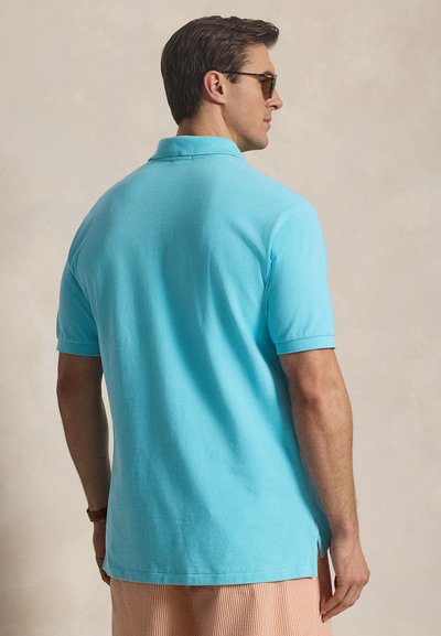 Άνδρας που φοράει τιρκουάζ πόλο μπλούζα με κοντά μανίκια, πορτοκαλί ριγωτά σορτς, γυαλιά ηλίου και ρολόι χειρός, φωτογραφημένος από πίσω σε ουδέτερο φόντο.