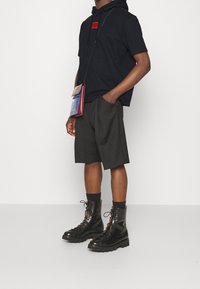 Schwarzer Kapuzenpullover mit einem roten Logo-Patch, graue Shorts und schwarze Schnürstiefel; lässiges Ensemble mit einer kleinen Umhängetasche.