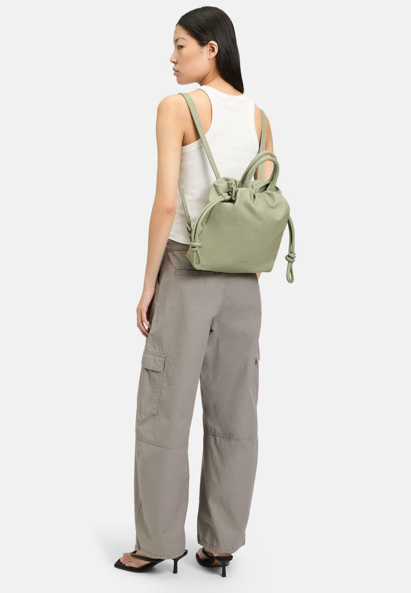 Sac à dos en cuir vert avec fermeture à cordon et poignées circulaires, porté sur un pantalon cargo gris, mettant en avant une texture lisse et un design minimal.
