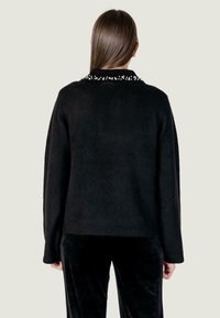 Jersey negro con un acabado texturizado y un cuello de cuentas, con un corte relajado y mangas largas, combinado con pantalones de terciopelo negro.