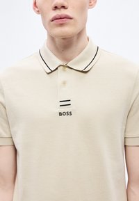 Μπεζ πόλο πουκάμισο από υφασμάτινο ύφασμα με υφή. Διαθέτει μαύρο περιθώριο στο γιακά, δύο μαύρες ρίγες και το λογότυπο "BOSS" κεντραρισμένο κάτω από τον γιακά.