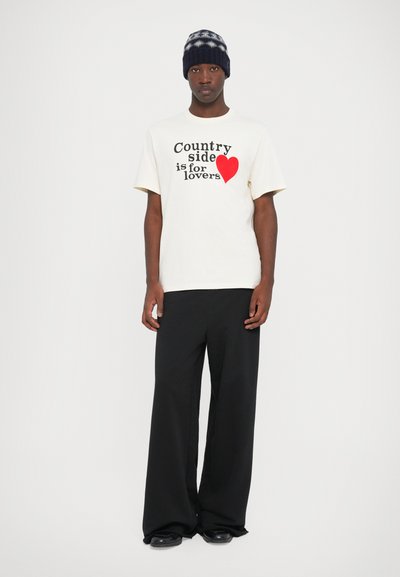 Cr�ème t-shirt met zwarte tekst en een rode hartgrafiek, gecombineerd met brede zwarte broek. Model draagt een gebreide muts met een patroon.