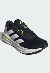 Czarne buty sportowe z siateczkowej cholewki, z białym logo w trzy paski, zielonymi akcentami i wyściełaną podeszwą. Charakteryzują się zaokrąglonym noskiem i sznurowanym zapięciem.