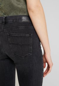 Primer plano de una persona que lleva unos jeans Diesel de color gris oscuro, mostrando el bolsillo trasero y la etiqueta del logo, con un brazo relajado a su lado.