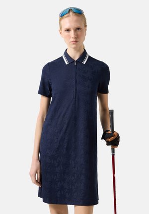 Kvinde i marineblå struktureret polokjole, der holder en golfkølle med handsker på, iført solbriller på hovedet og stående mod en ensfarvet baggrund.