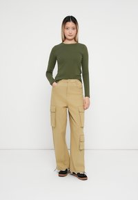 Un top cu mânecă lungă de culoare verde măslinie, asortat cu pantaloni cargo bej, care au multiple buzunare. Modelul poartă pantofi negri cu accente albe.