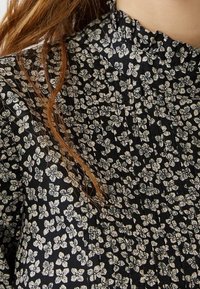 Mustakankainen kangas, jossa on pieni valkoinen kukkakuosi ja laskoksia, näkyy osa henkilön olkapäästä ja punaruskea hiussuortuva.