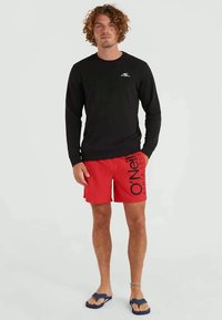 Juodas ilgomis rankovėmis marškinėliai, raudoni maudymosi šortai su dideliu juodu prekės ženklo logotipu ir mėlynos šlepetės. Kasdienė vasaros apranga, demonstruojanti ryškias spalvas.