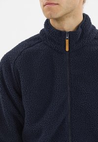 Laivaston fleece-takki, jossa on tekstiilinäytteen muotoinen, pörröinen pinta, puoliveto-kaulus ja kontrastivärinen musta vetskari; vetskarin vetimessä on pieni vaaleanruskea yksityiskohta.