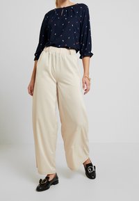 Námořnický top s barevnými vzory listů, béžové kalhoty s elastickým pasem a širokými nohavicemi a černé mokasíny s kulatými akcenty.