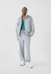 Giacca in pile grigia chiara con colletto alto, t-shirt teal sotto e pantaloni della tuta grigi coordinati. Accentuata da polsini elasticizzati e vita regolabile con cordino.