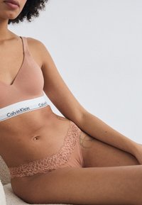 Bralette en coton rose clair avec une large bande blanche portant le logo "Calvin Klein". Sous-vêtements roses transparents avec des bordures en dentelle assorties. Textures lisses.