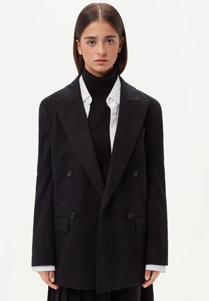 Schwarzer zweireihiger Blazer aus Wollmischung mit Reverskragen, zwei vorderen Taschen und Knopfdetails. Getragen über einem schwarzen Rollkragenpullover.
