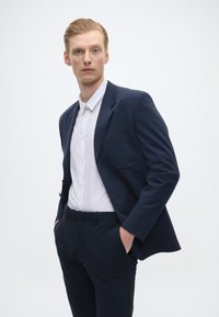 Blazer azul-marinho com um único bolso, camisa branca por baixo e calças azul-marinho a condizer. Tecido suave com um corte ajustado.