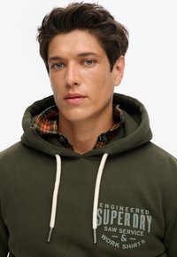 Olijfgroene hoodie met witte trekkoorden, voorzien van een vooraanstaand logo in lichtblauw. Daaronder een geruit flanellen kraag in oranje en zwart.