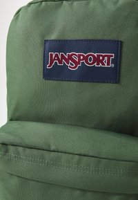 Zöld szövet hátizsák kék Jansport címkével. A felülete textúrás, és a hátsó zseben látható varrásokkal rendelkezik.