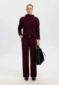 Mujer vestida con un suéter de canalé burdeos y pantalones de pierna ancha a juego, llevando un gran bolso de cuero negro, de pie contra un fondo sencillo.