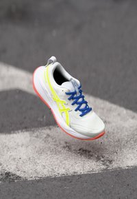 Chaussure de course avec une tige en mesh blanc, des accents jaune néon, des lacets bleus et une semelle en caoutchouc rose, surélevée au-dessus d'une bande de bitume.