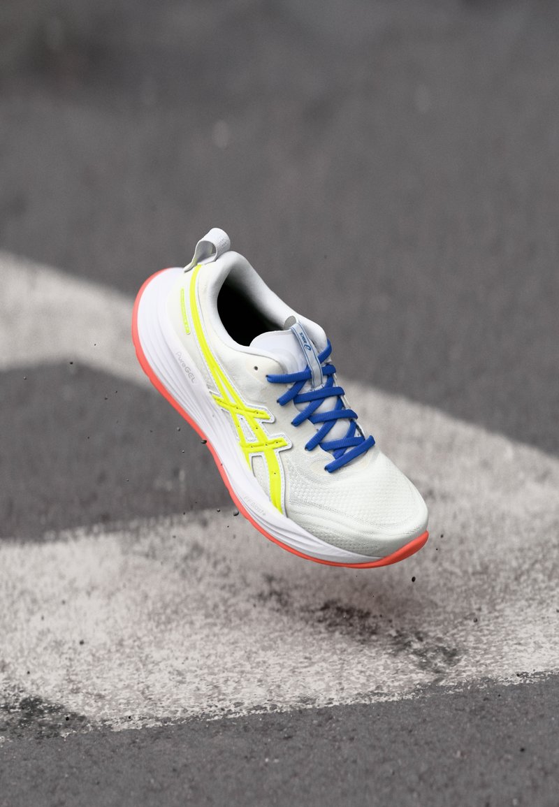 Chaussure de course avec une tige en mesh blanc, des accents jaune néon, des lacets bleus et une semelle en caoutchouc rose, surélevée au-dessus d'une bande de bitume.