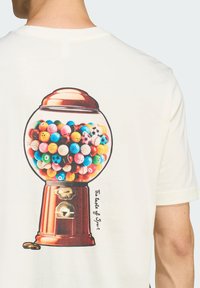 Krémové tričko s farebnou grafikou žuvačkovej gule, ktorá je naplnená rôznymi vzorovanými guľami, na jednoduchom pozadí.