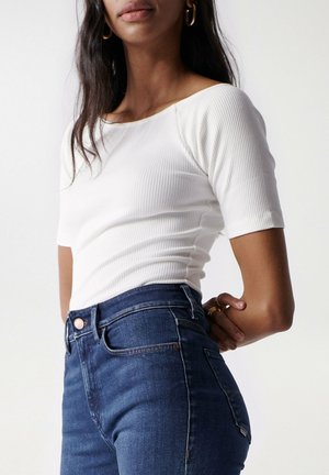 Frau trägt weißes, geripptes kurzärmeliges Oberteil und hoch taillierte blaue Jeans, mit goldenen Creolen und Ringen an der linken Hand.