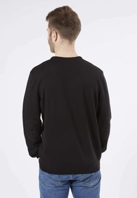 Un sweat noir à col rond, avec des poignets et un ourlet côtelés, présentant des pièces de renfort aux coudes. Porté avec un jean bleu, vu de dos sur un fond blanc.