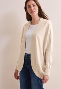 Beige cardigan gemaakt van zacht materiaal, met een geribbelde open voorkant en manchetten, ontspannen pasvorm, en gecombineerd met een witte top en blauwe jeans.