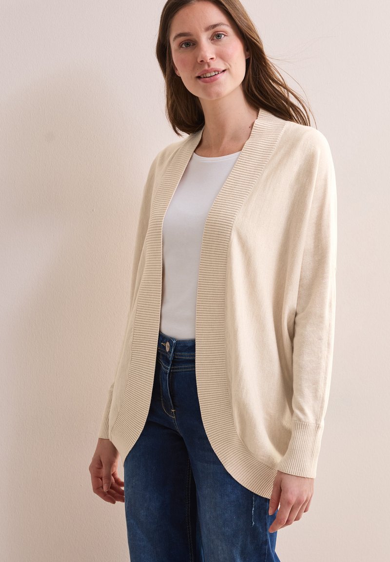 Beige cardigan gemaakt van zacht materiaal, met een geribbelde open voorkant en manchetten, ontspannen pasvorm, en gecombineerd met een witte top en blauwe jeans.
