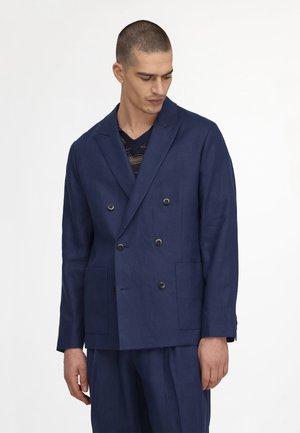 Mann trägt marineblauen, doppelt geknöpften Blazer mit schwarzen Knöpfen und passende marineblaue Hose, sieht nach unten zur Seite.